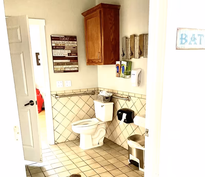 A clean and accessible bathroom with beige tiled floor and walls. The bathroom features a white toilet with safety grab bars on both sides, a wooden cabinet mounted on the wall above the toilet, a small waste bin, a soap dispenser, and a wall-mounted paper towel holder. There is a decorative sign on the wall with inspirational text and a wooden coat rack with hooks. The door to the bathroom is open, showing a glimpse of an adjacent room with a red bedspread.