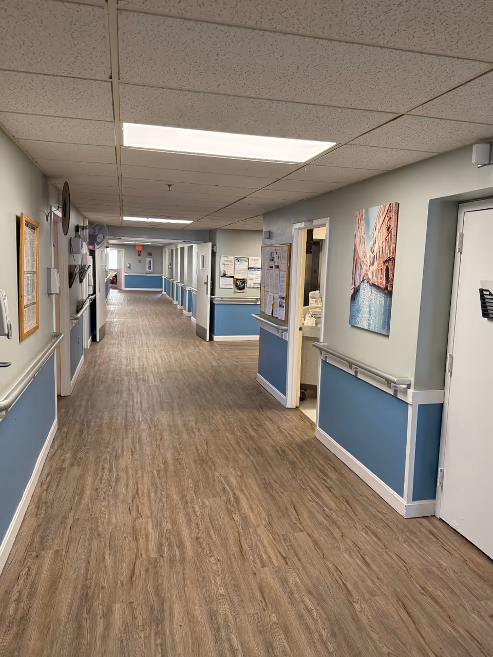 A long, clean hallway in a nursing and rehabilitation facility with wood-patterned flooring, light blue and white walls, handrails on both sides, fluorescent ceiling lights, bulletin boards, and a framed picture of a canal scene on the right wall.