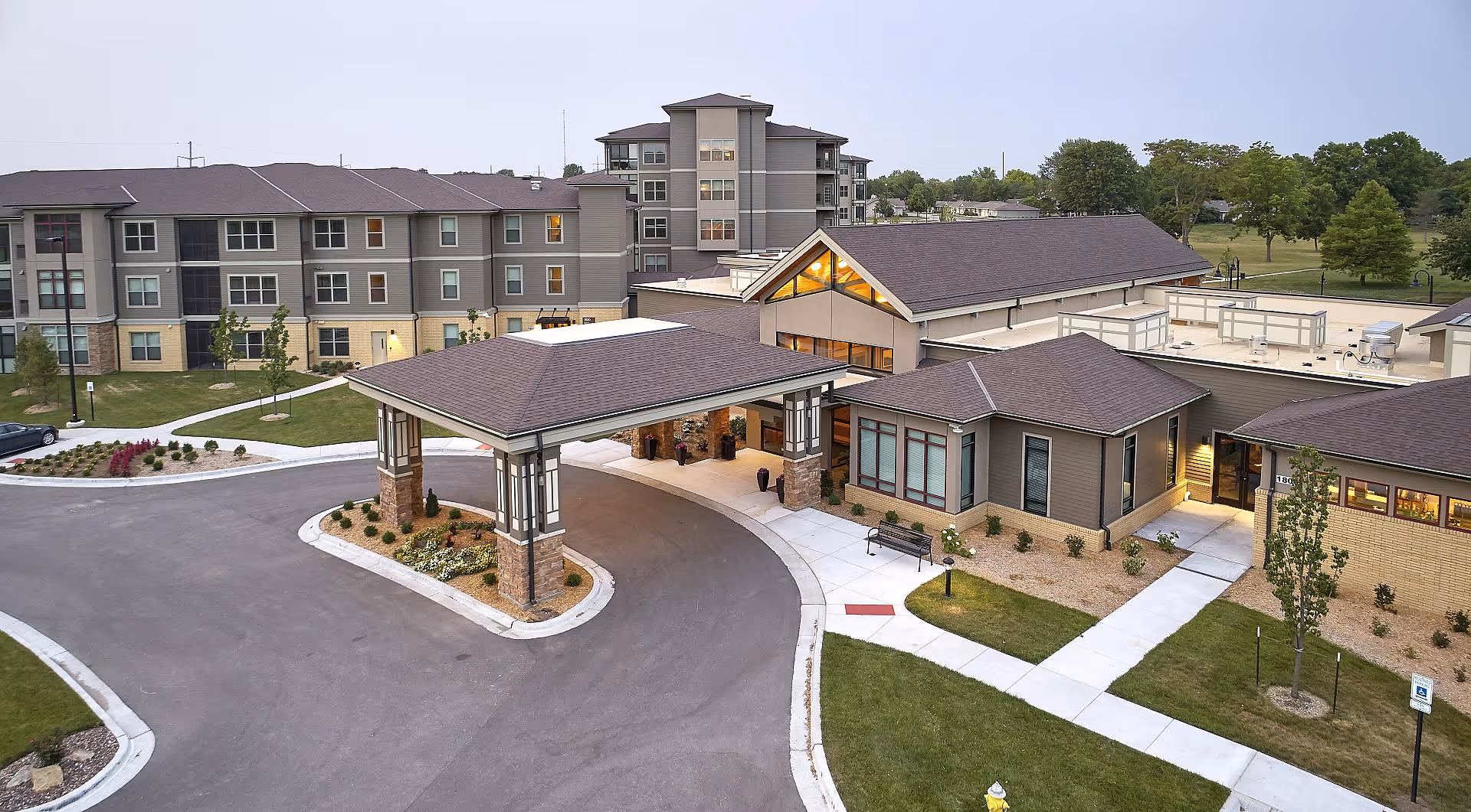 Aerial view of a senior living campus showing the main entrance with a porte-cochère, multi-story residential buildings, driveways, and landscaped grounds.
