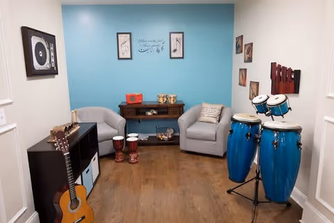 A cozy music-themed room with two gray armchairs, a wooden console table with small drums and a red radio, a black shelving unit with a guitar leaning against it, two small hand drums on the floor, and a set of blue conga drums on a stand. The walls are decorated with framed pictures and a musical quote on a blue accent wall.