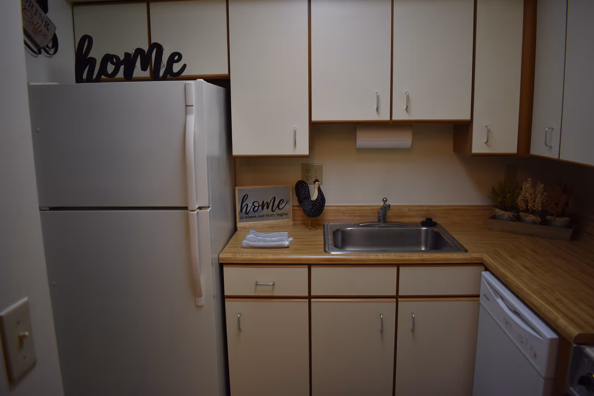 A compact kitchen featuring a white refrigerator, white cabinets with silver handles, a stainless steel sink, and a wooden countertop. Decorative items include a sign that says 'home is where our story begins,' a black and white polka dot oven mitt hanging on the wall, and a tray with small plants on the right side of the counter.