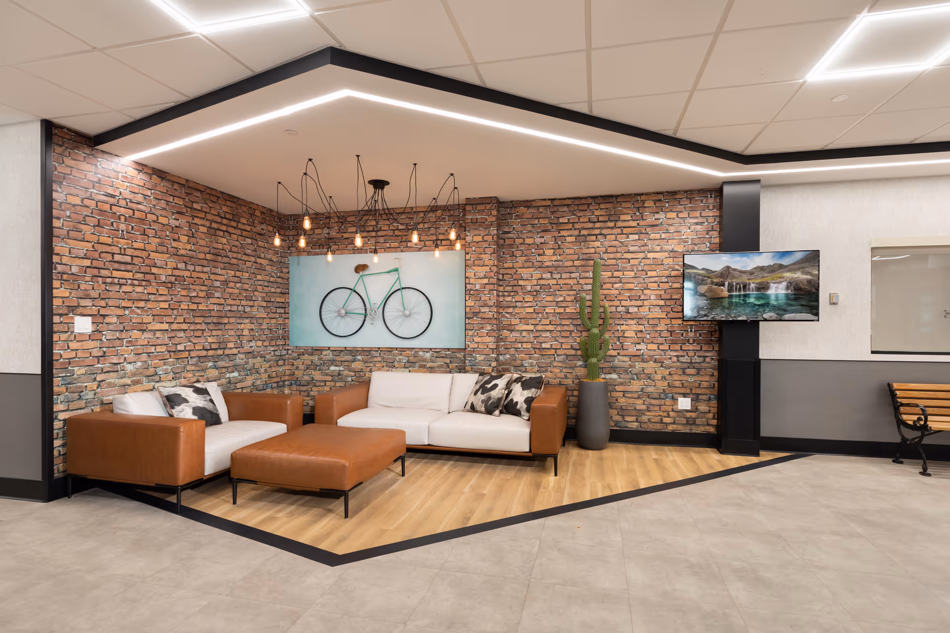 A modern lounge area with a white and brown leather sofa, matching ottoman, and a brown leather armchair. The walls are decorated with exposed brick and a large artwork of a green bicycle. A tall cactus plant in a gray pot stands next to a wall-mounted flat-screen TV displaying a scenic landscape. The ceiling features a unique light fixture with exposed bulbs and a geometric LED strip light. The floor transitions from light wood in the seating area to gray tiles in the surrounding space.