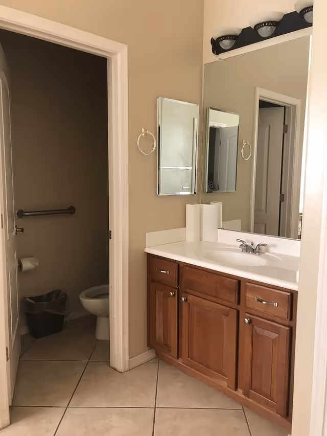 Bathroom vanity with sink, mirror and wooden cabinets next to a small toilet room with a grab bar.