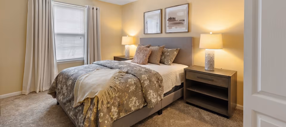 Well-lit furnished bedroom with a bed, patterned bedding, two nightstands with lamps, a window with curtains, and framed art above the headboard.