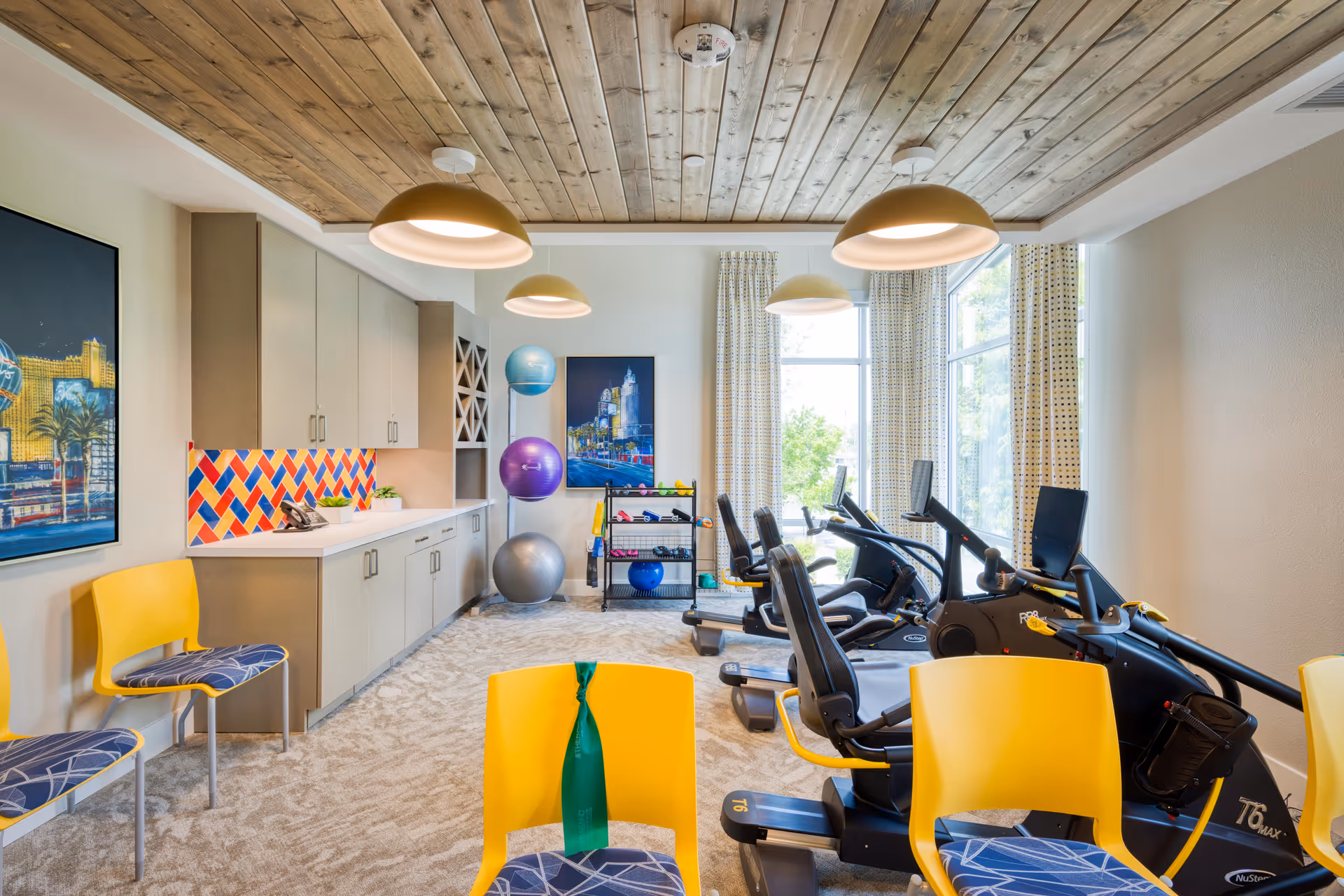 Bright fitness/activity room with recumbent exercise bikes, colorful chairs, storage cabinets and large windows.