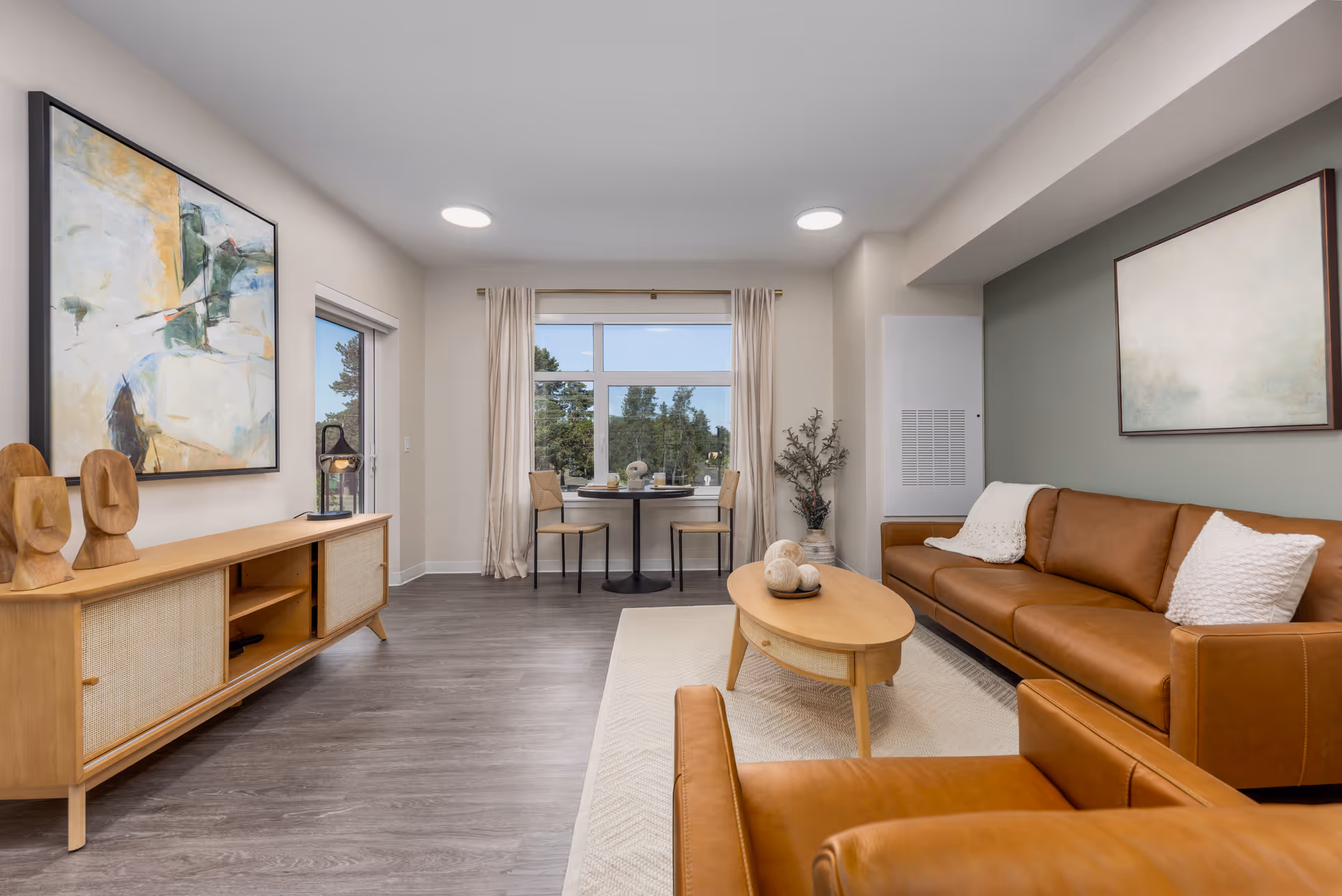 A modern living room with a brown leather sofa and matching armchair, a wooden coffee table with decorative spheres, a wooden sideboard with two wooden sculptures and a lantern, abstract paintings on the walls, and a small dining table with two chairs near a large window with beige curtains showing trees outside.