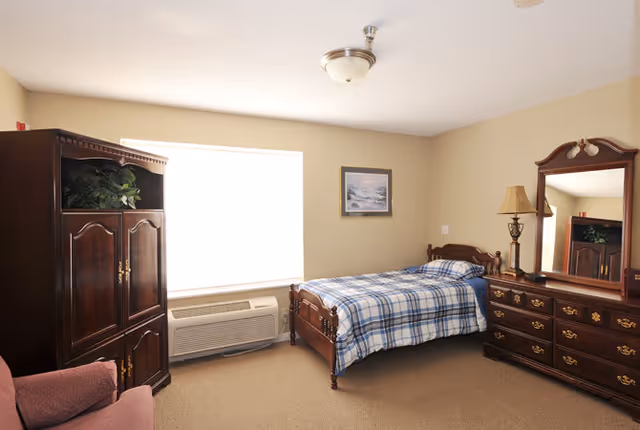 A cozy bedroom with a single bed covered in a blue and white plaid blanket, a wooden dresser with a large mirror, a wooden armoire with a plant on top, a pink upholstered chair, and a window with an air conditioning unit below it. The walls are beige and there is a framed picture hanging above the bed.