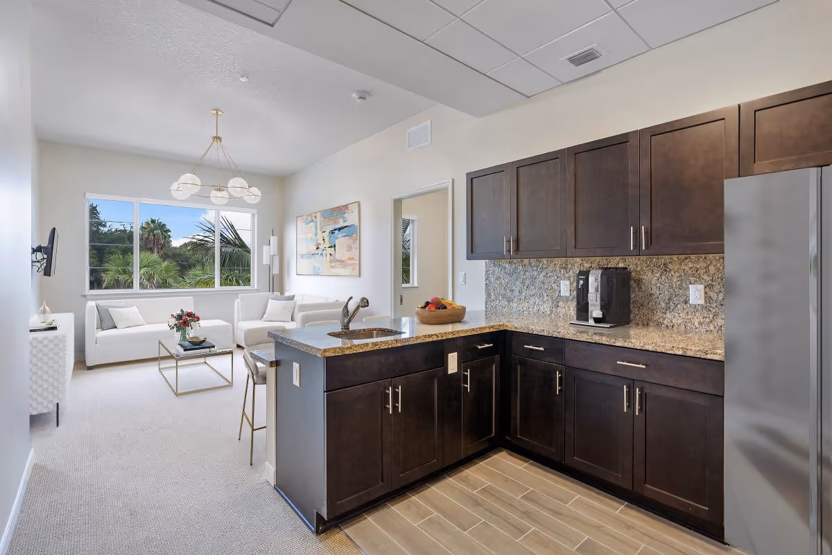 Modern kitchen with dark wood cabinets, granite countertops, a coffee maker, and a stainless steel refrigerator. The kitchen opens into a bright living room with white sofas, a glass coffee table, a wall-mounted TV, and a large window showing palm trees outside.