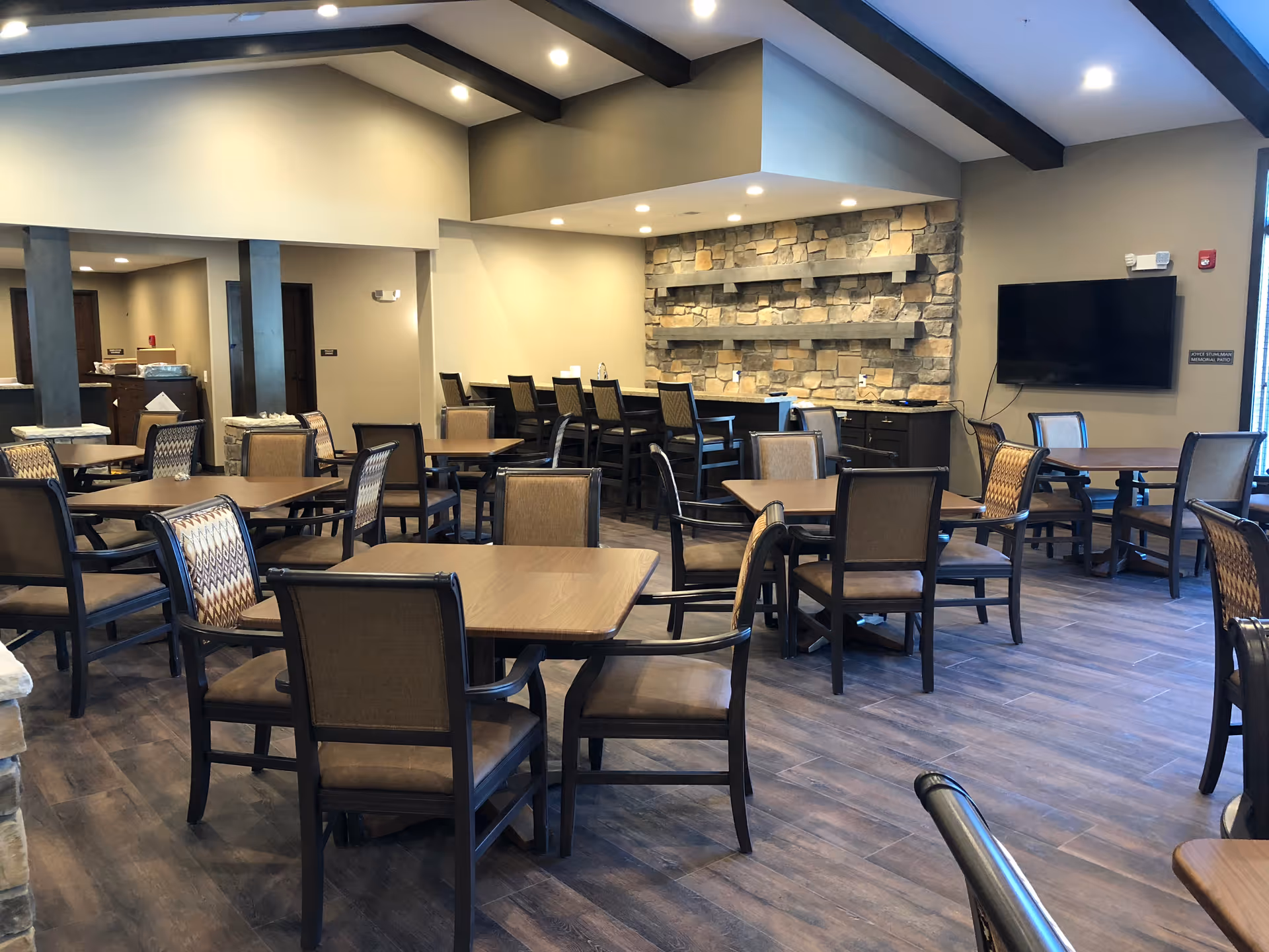 Spacious communal dining room with multiple tables and chairs, a stone accent wall, counter seating and a wall-mounted TV.