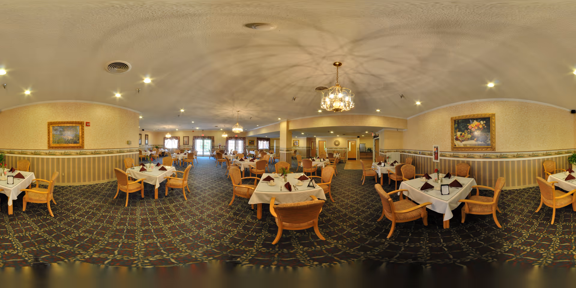 A spacious dining room with multiple tables covered in white tablecloths and set with napkins, glasses, and menus. The room features patterned carpet, beige walls with decorative wainscoting, framed paintings, and chandeliers hanging from the ceiling. Wooden chairs with cushions surround the tables, and large windows at the far end allow natural light to enter.