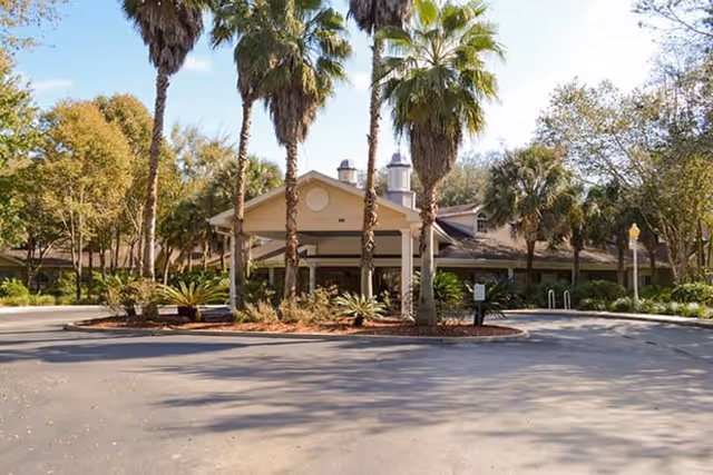 Exterior view of a single-story building with a covered entrance surrounded by palm trees and other greenery under a clear blue sky.