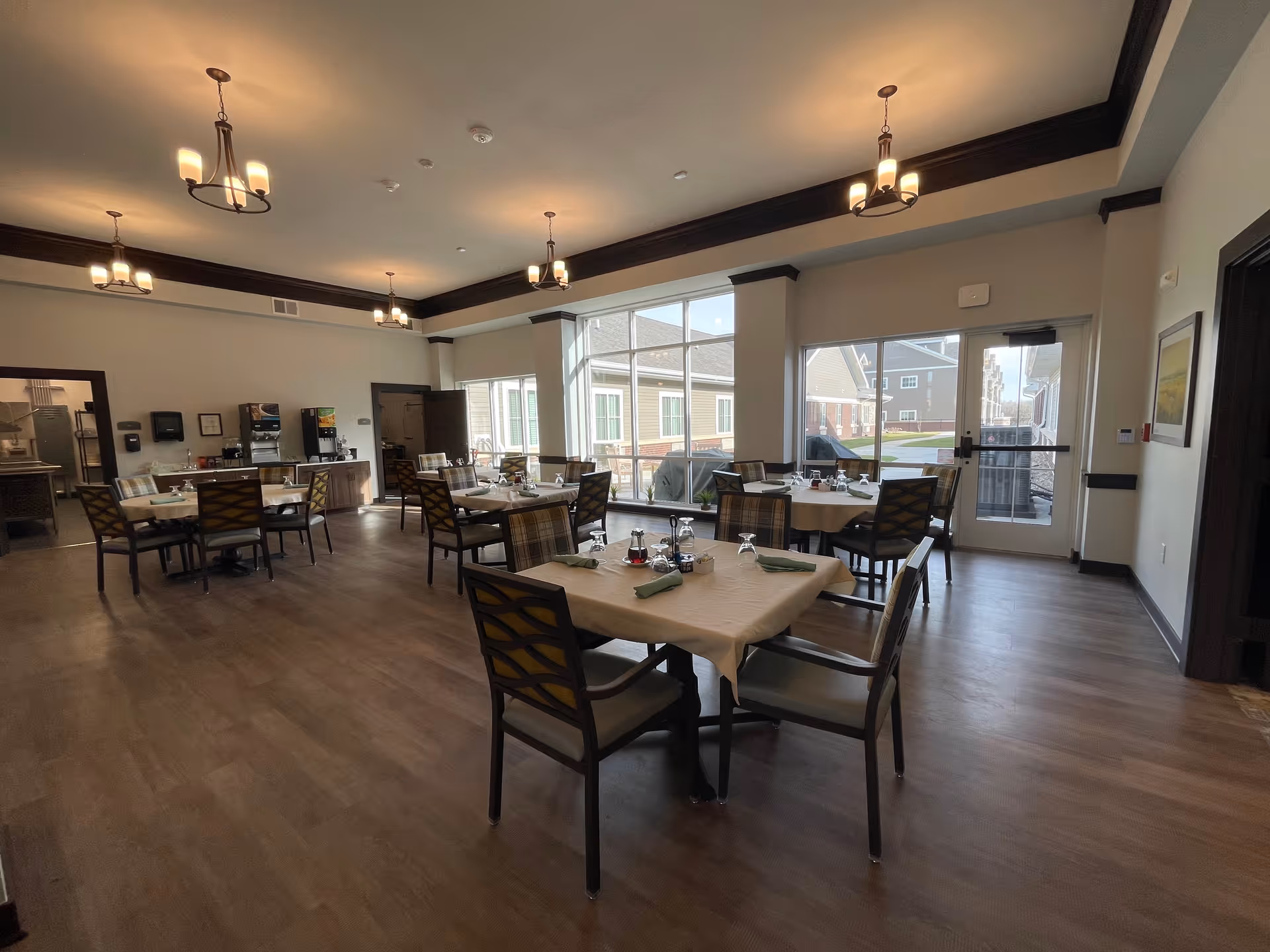 A spacious dining room with several tables covered with beige tablecloths, each set with napkins, glasses, and condiments. The room has large windows letting in natural light, wooden flooring, and multiple ceiling light fixtures. There is a beverage station along one wall and a door leading outside.