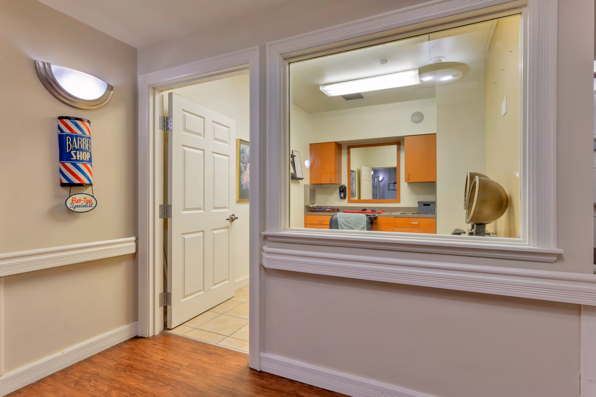 Interior view of a hallway in a senior living facility with a white door open leading to a room with tiled floor. On the left wall, there is a decorative barber shop sign with red, white, and blue stripes and the words 'Barber Shop' and 'Flat-Top Specialist'. To the right, there is a large window looking into a room with wooden cabinets, a mirror, and a vintage hair dryer chair.