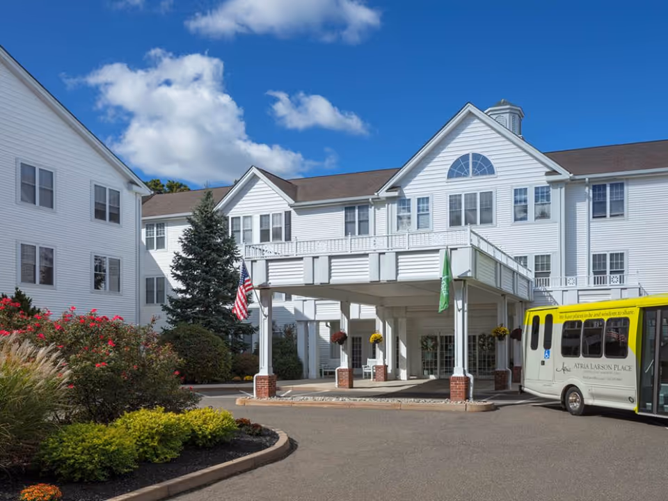 Exterior view of Atria Larson Place, a white senior living building with covered entrance and bus.
