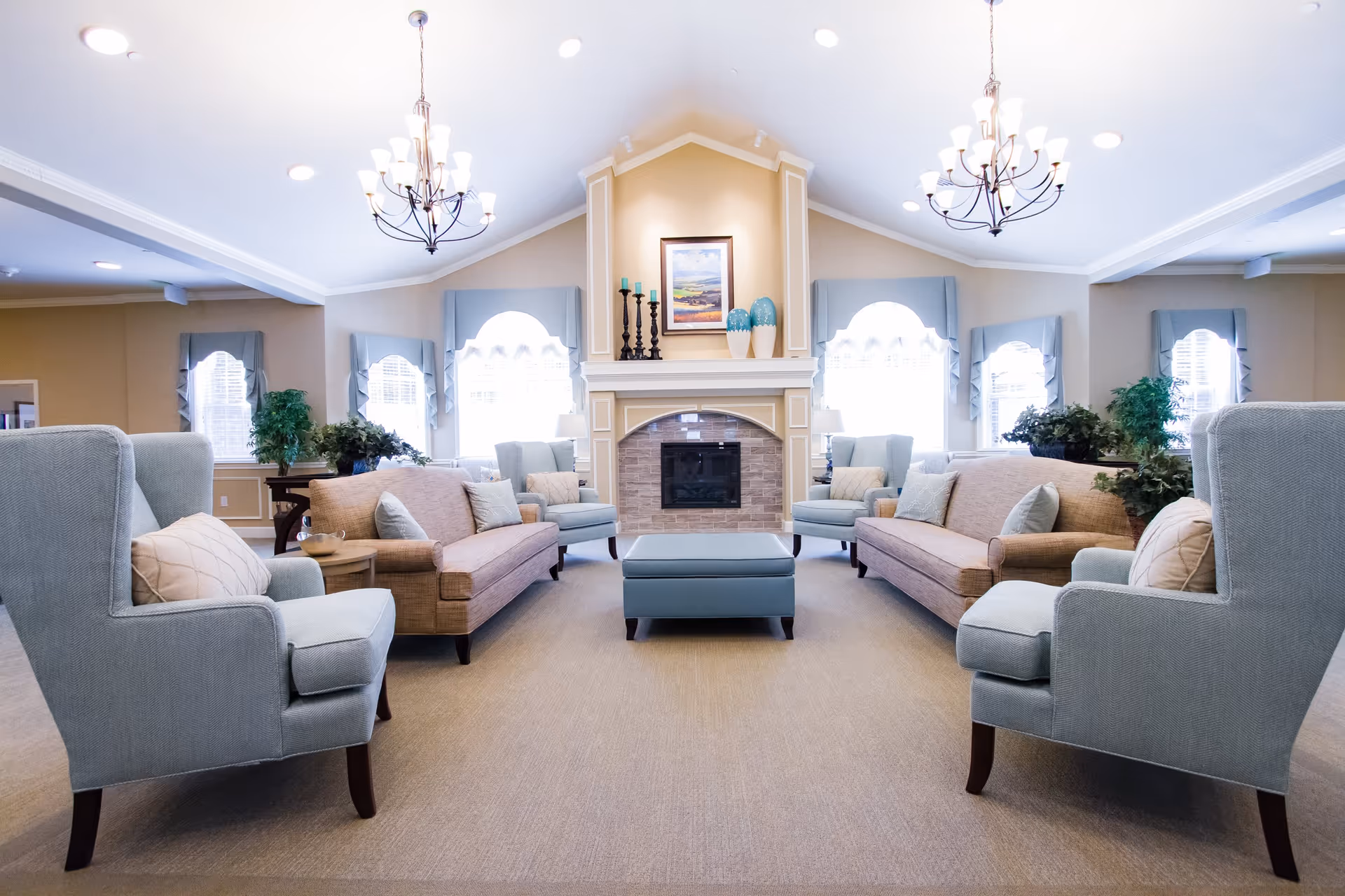 Spacious communal living room with sofas, armchairs, a central ottoman and a fireplace beneath chandeliers.