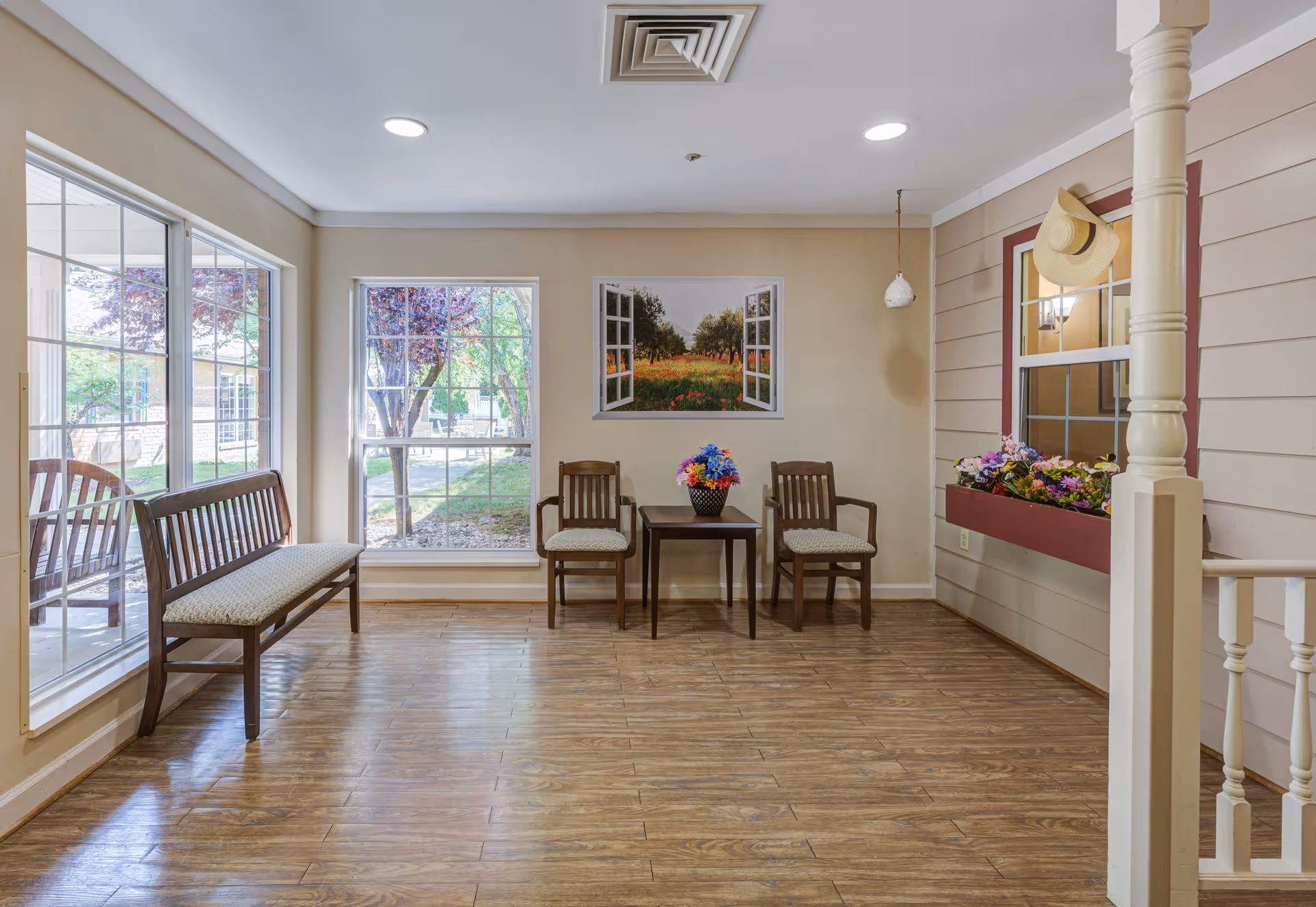 A bright sitting area with wooden floor and beige walls. There are two wooden chairs with cushions and a small table with a colorful flower arrangement between them. To the left, a wooden bench with a cushion is placed near large windows showing an outdoor garden. On the wall above the chairs, there is a framed picture of an open window looking out onto a field with trees and flowers. A window box with flowers and a hanging hat is on the right wall. The ceiling has recessed lighting and an air vent.