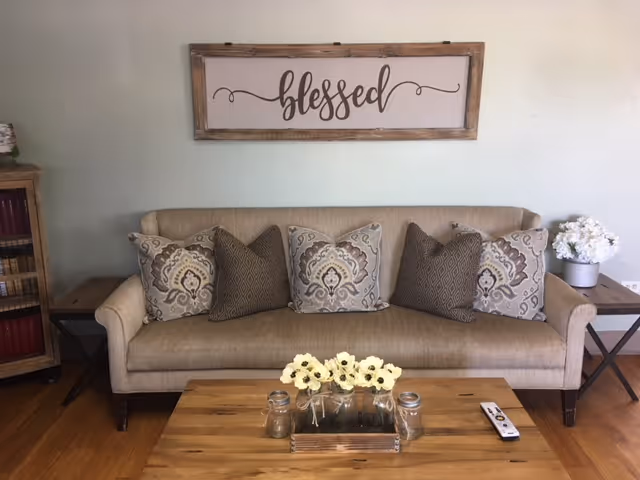 A beige sofa with five decorative pillows in patterned and solid designs is placed against a light-colored wall. Above the sofa, a wooden framed sign with the word 'blessed' in cursive hangs on the wall. On either side of the sofa are wooden side tables, one with a white flower arrangement. In front of the sofa is a wooden coffee table with a tray holding three small jars and a bunch of white flowers, along with a TV remote control. The floor is wooden.