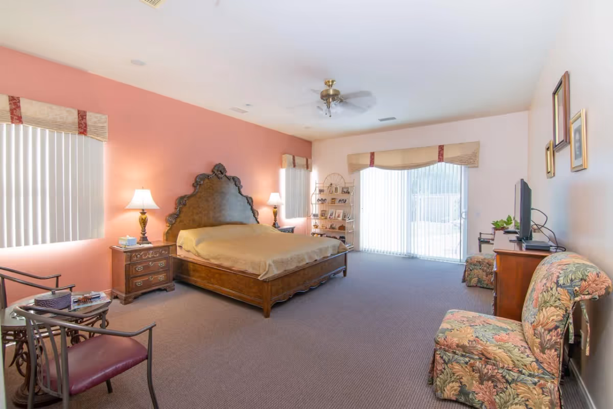 A spacious bedroom with a large wooden bed featuring an ornate headboard, two bedside tables with lamps, a floral upholstered chair, a small table with two chairs, a TV on a wooden dresser, and large windows with vertical blinds and valances letting in natural light.