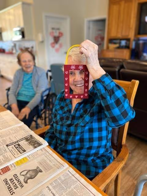 An elderly woman in a blue and black checkered shirt is sitting at a table holding a red paper frame decorated with hearts in front of her face, smiling. Another elderly woman is sitting in the background, smiling. The setting appears to be a cozy indoor common area with wooden furniture and kitchen cabinets visible.