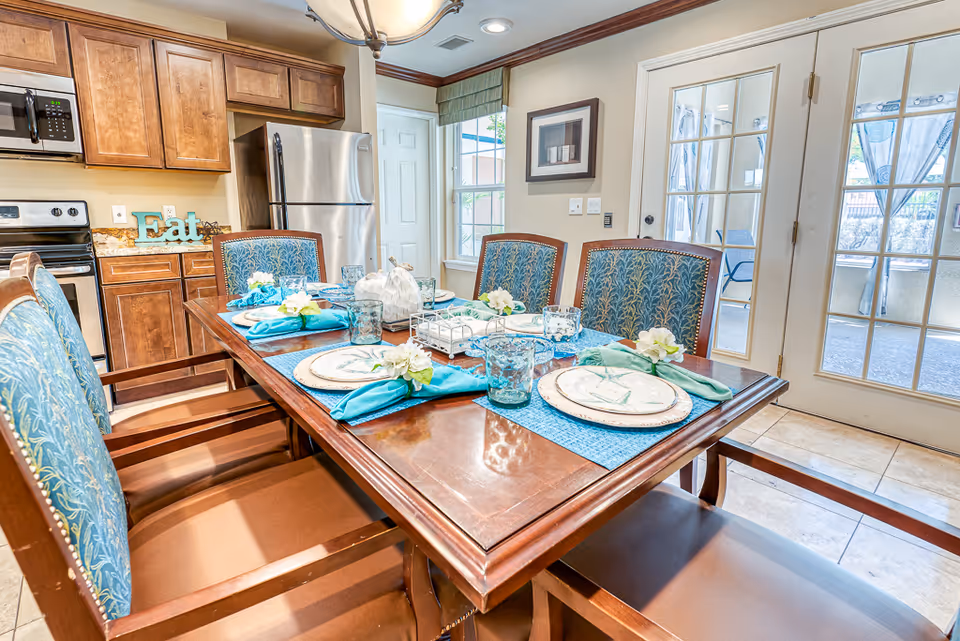 Dining table set with place settings and blue-green accents in a bright kitchen-dining area with wooden cabinets and glass patio doors.