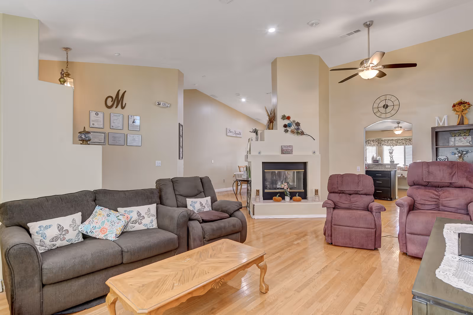 A cozy living room with a gray sofa and two purple recliners arranged around a wooden coffee table. The room features a light wood floor, a ceiling fan with lights, and a fireplace decorated with small pumpkins and flowers. The walls are painted beige, and there are decorative items including wall art, a clock, and framed certificates on the walls.