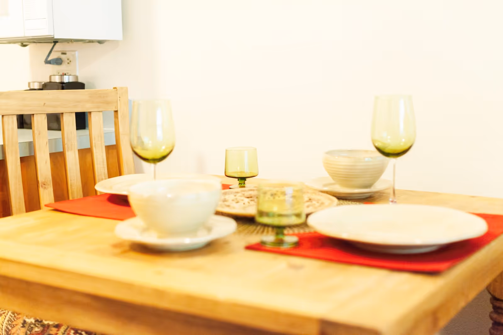 Wooden dining table set with plates, bowls and green-stemmed glasses with a wooden chair and kitchen counter in the background.