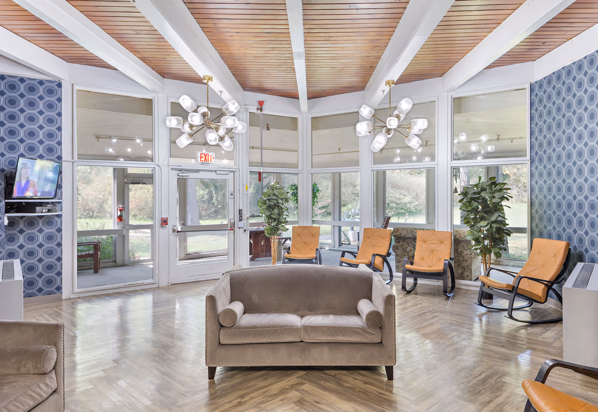 A bright and spacious living room area with a wooden ceiling and large windows letting in natural light. The room features a gray sofa in the center, several orange cushioned chairs arranged around the perimeter, two modern chandeliers, blue patterned wallpaper on the side walls, and a wall-mounted TV on the left. There are also two potted plants near the windows and a glass exit door leading outside.