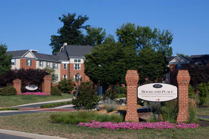 Exterior view of Rockland Place, a senior living and memory care community, showing a brick entrance sign surrounded by pink flowers and greenery, with a multi-story brick building and trees in the background under a clear blue sky.