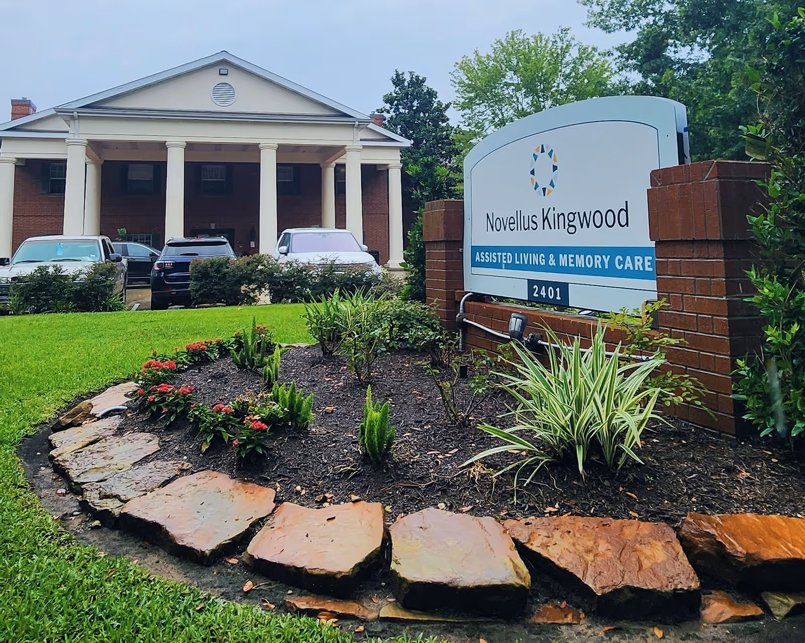 Entrance sign reading 'Novellus Kingwood' in front of the facility's columned brick facade with a landscaped flowerbed and parked cars.