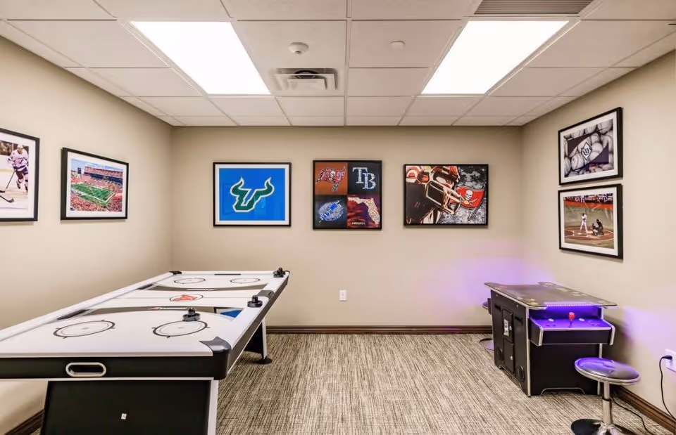 A game room with an air hockey table and an arcade game machine. The walls are decorated with framed sports-themed pictures including hockey, football, and baseball images. The room has beige walls, a carpeted floor, and a drop ceiling with fluorescent lighting.