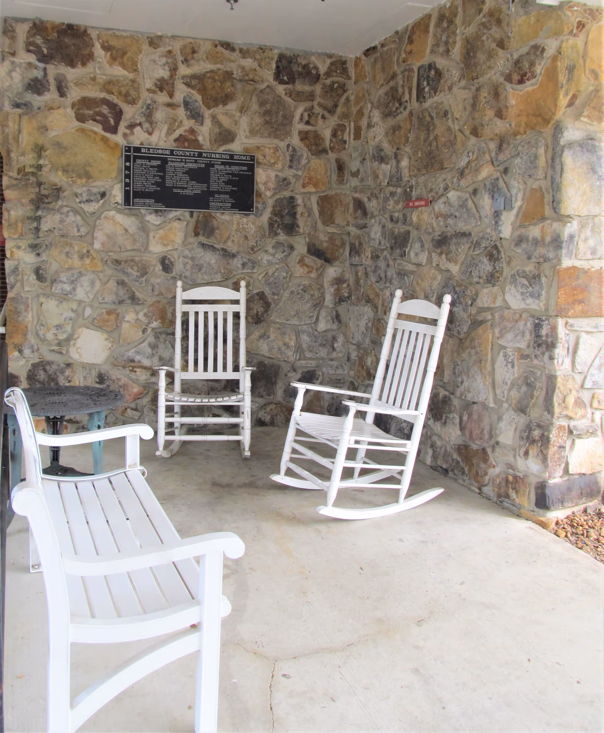 A covered outdoor seating area with a stone wall background featuring two white wooden rocking chairs, a white wooden bench, and a small round metal table. A plaque is mounted on the stone wall, and there is a small red sign on the right side of the wall.