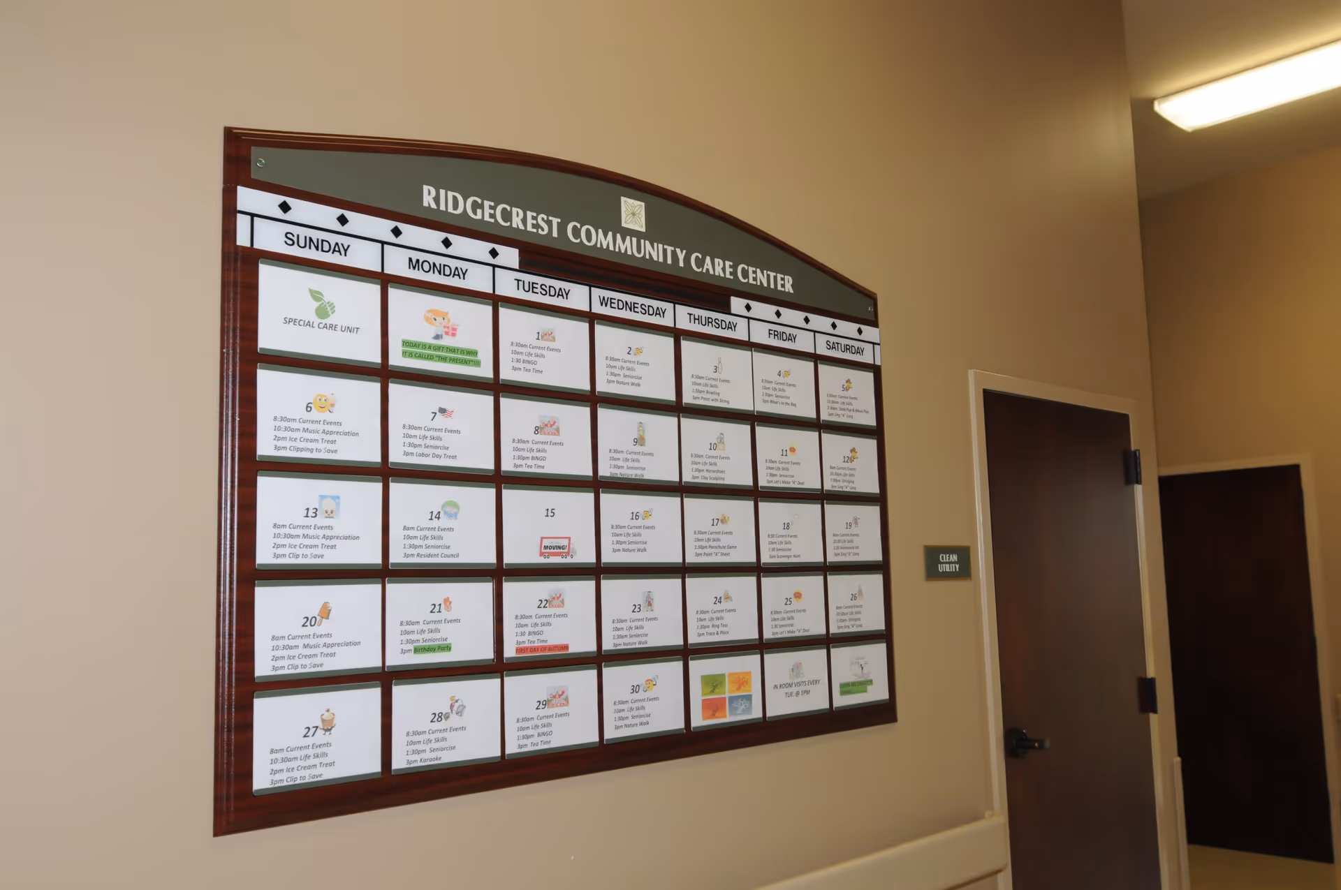 A wall-mounted monthly activity calendar for Ridgecrest Community Care Center, showing daily events and activities. The calendar is framed in dark wood and mounted on a beige wall next to two closed brown doors, one labeled 'Clean Utility'.