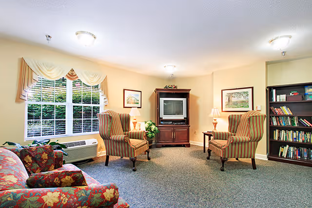 Cozy lounge with patterned armchairs and a floral sofa facing a TV cabinet, bookcase, lamps, and a large window.