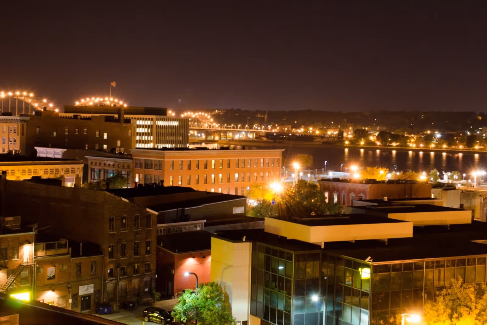 Nighttime cityscape featuring illuminated buildings, a river reflecting lights, and a brightly lit bridge in the background.