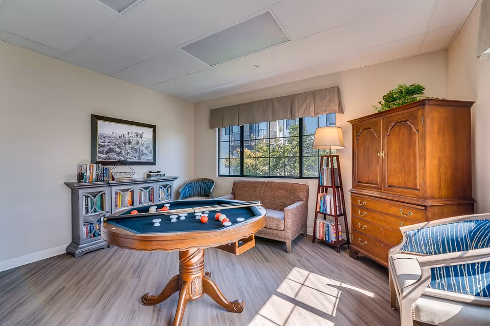 A bright and cozy senior living common room with a small pool table in the center. The room features a wooden cabinet, a bookshelf with books, a floor lamp, a brown sofa, and two armchairs. A large window lets in natural light, illuminating the wood-style flooring and neutral-colored walls.