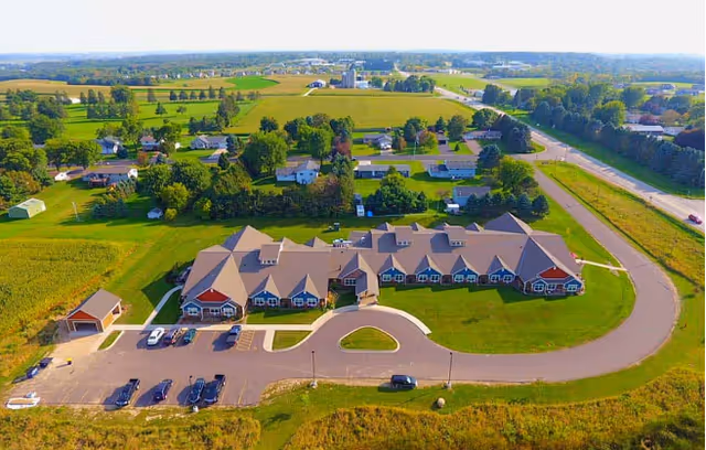 Aerial view of a long single-story senior living building with a circular driveway, parking lot, and surrounding farmland.