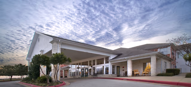 Front entrance of a senior living building with a covered porte-cochere, driveway, rocking chairs on the porch, and landscaped trees under a cloudy sky.