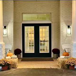 Front entrance with double glass doors framed by pillars, lit wall sconces, and autumn decorations including pumpkins, mums, and hay bales.