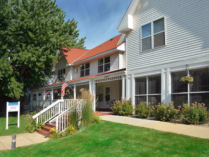 Exterior view of Elder Homestead, a two-story white building with a red roof, surrounded by green grass, bushes, and trees. There is a white railing along the front porch with steps leading up to the entrance, and an American flag is displayed near the door. A sign with the facility name is visible on the lawn.