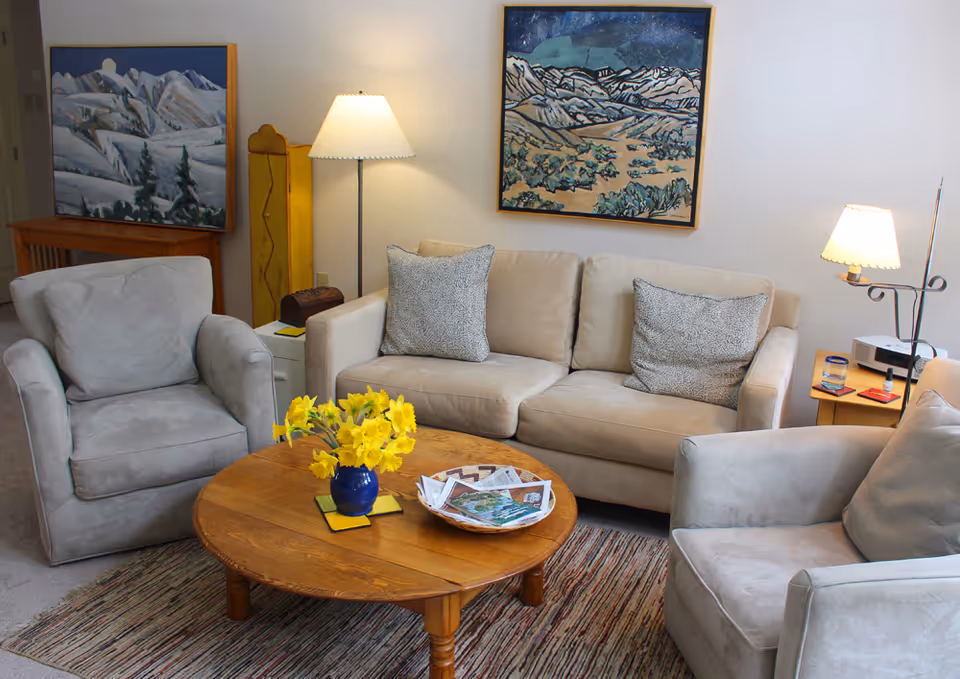 Cozy living room with a beige sofa and two armchairs around a round wooden coffee table topped with a blue vase of yellow flowers and magazines.