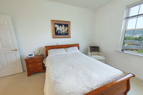A bright bedroom with a wooden bed frame and white bedding, a wooden nightstand with a phone and a clock, a cushioned armchair with a decorative throw, a window showing an outdoor view, and a framed artwork on the wall above the bed.