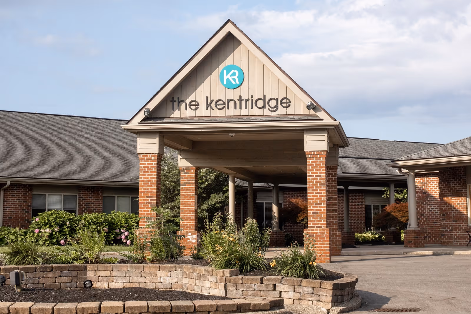 Covered brick entrance and gabled portico of The KentRidge senior living building with landscaped planters in front.