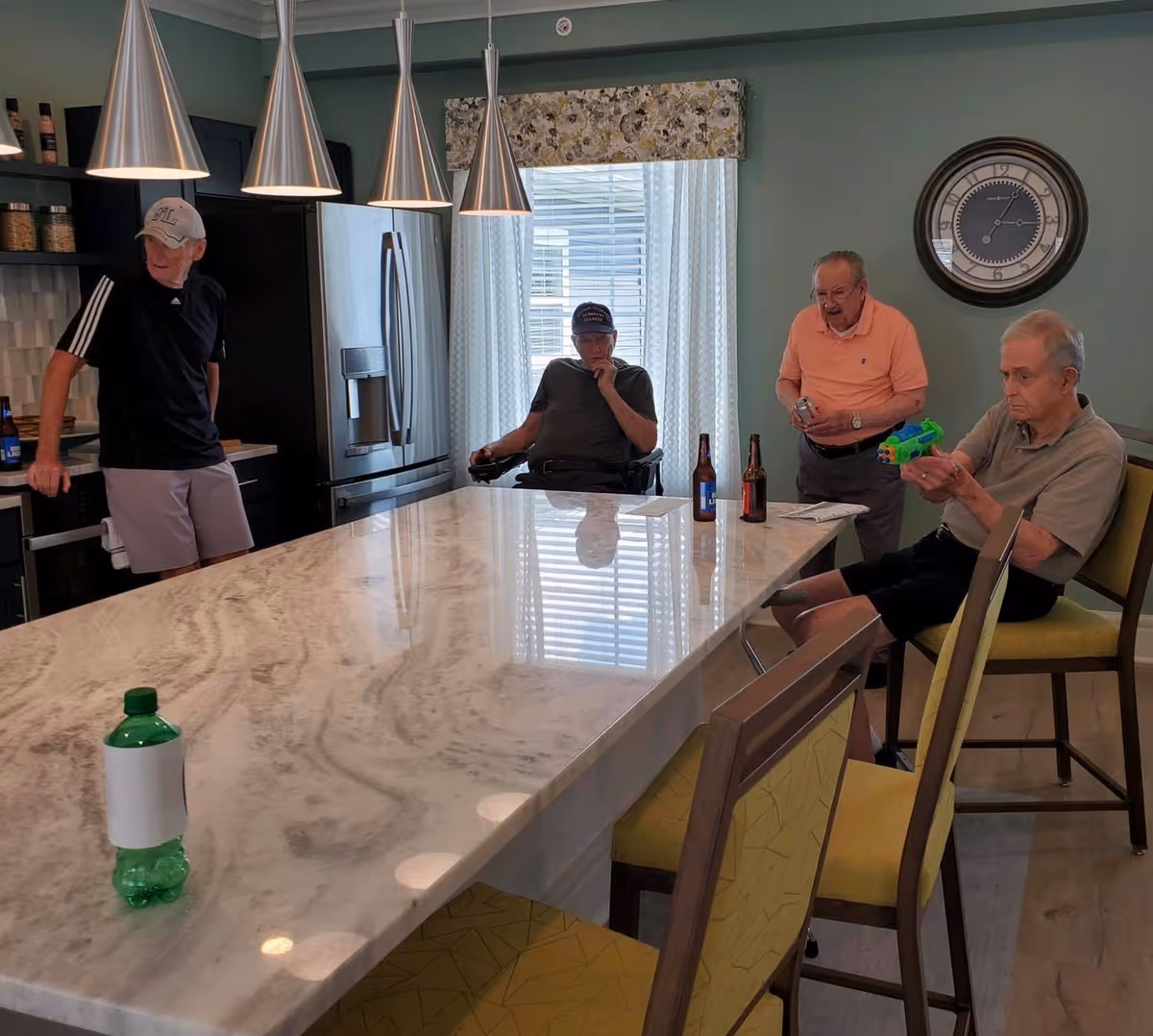 Four elderly men in a modern kitchen area with a large marble island countertop. One man is standing near the stove, another is seated in a wheelchair near the window, a third man is standing holding a can, and the fourth man is seated holding a toy water gun. The kitchen has stainless steel appliances, pendant lights, and a large wall clock.