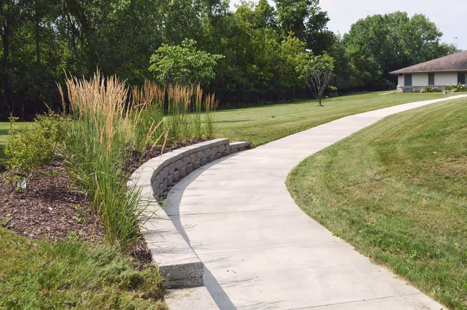Curved concrete walkway through a landscaped lawn with ornamental grasses and trees leading toward a low building.
