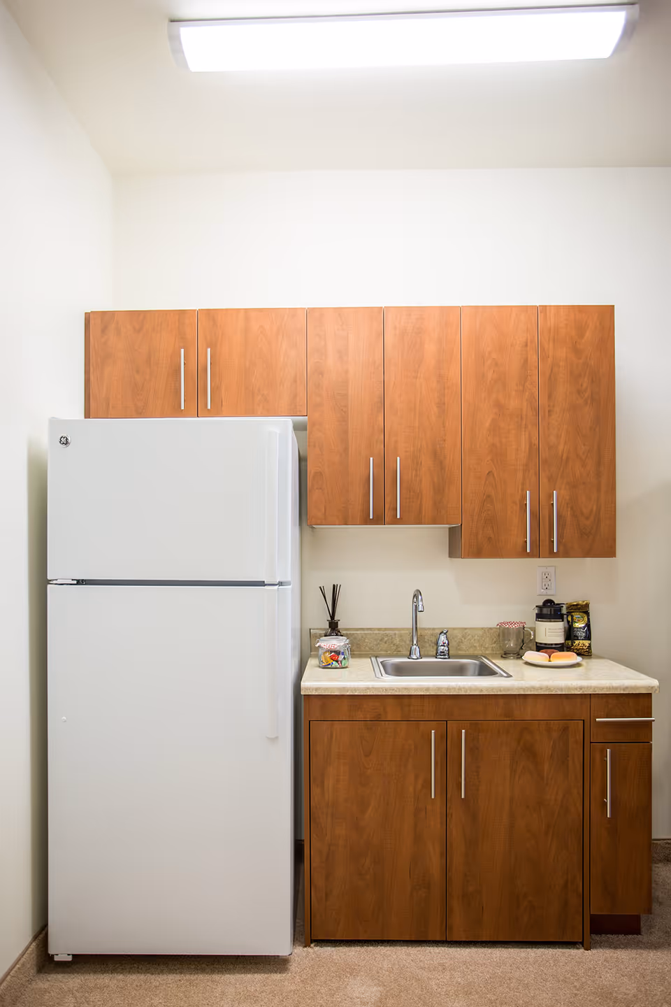 A small kitchen area with a white refrigerator on the left and wooden cabinets above and below a countertop with a sink. On the countertop, there are a few items including a glass jar with candy, a reed diffuser, a glass mug, a coffee container, and a plate with two donuts.