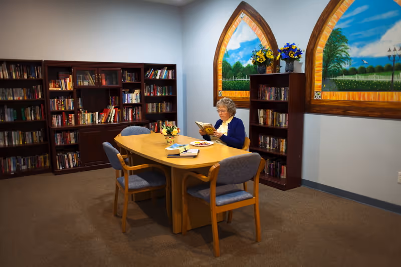 An elderly woman sitting at a wooden table in a library or reading room, reading a book. The room has bookshelves filled with books, two decorative wall murals resembling arched windows showing outdoor scenes, and a vase with flowers on the table.