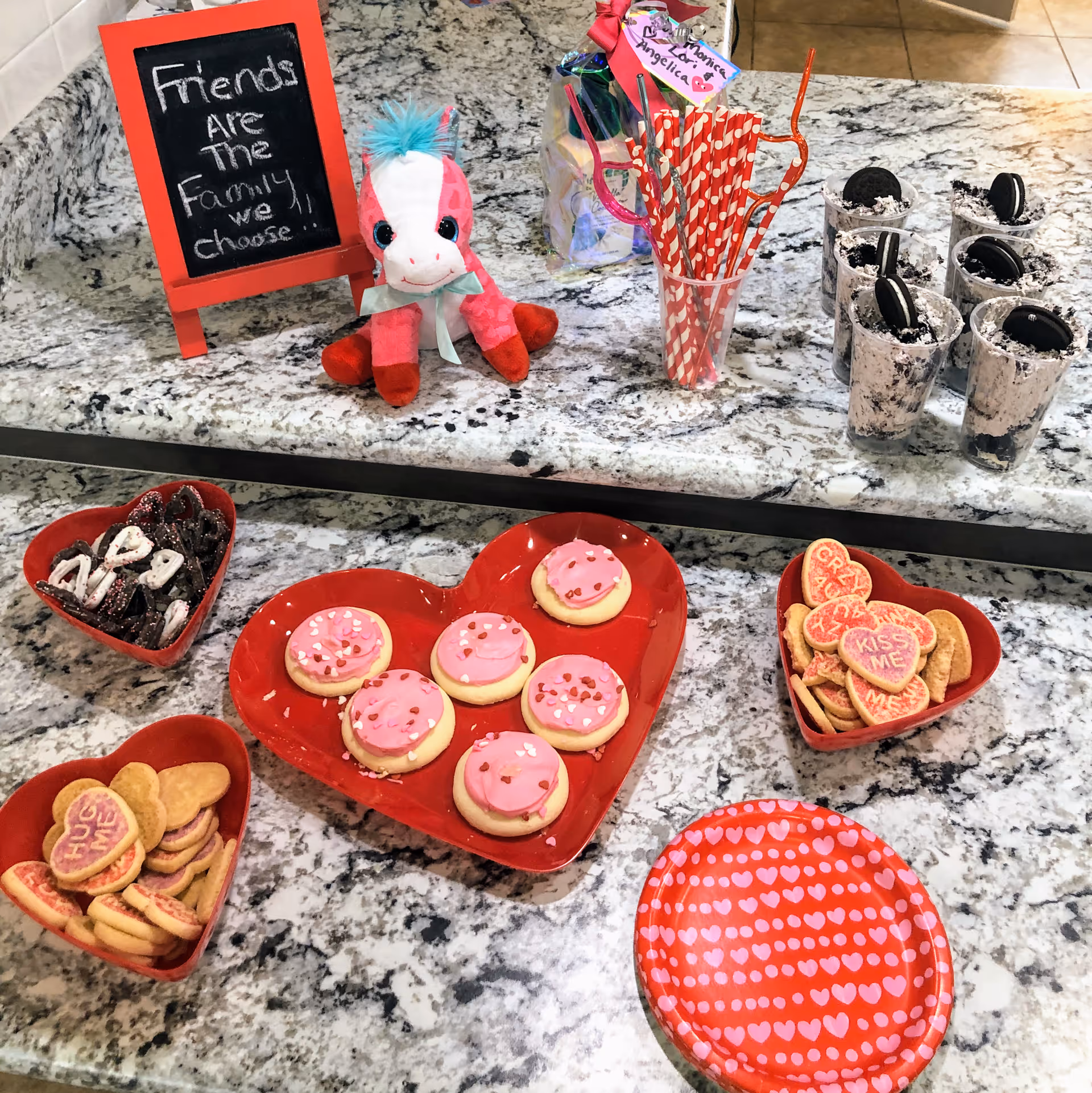 A countertop with heart-shaped red plates filled with decorated cookies, including pink frosted cookies and heart-shaped cookies with messages like 'HUG ME' and 'KISS ME'. There are also small cups filled with cookies and cream dessert topped with Oreo cookies, a cup with red and white striped straws, a small stuffed pink and white unicorn toy, and a small chalkboard sign that reads 'Friends are the family we choose'.