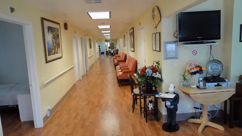 A long hallway in a senior living facility with wooden flooring and pale yellow walls. Several orange armchairs are lined up along the right side of the hallway. There are framed pictures on the walls and handrails on the left side. A small table with a flower arrangement and a statue of a waiter holding a tray is near a mounted TV on the right. At the far end of the hallway, a person in a wheelchair is visible.