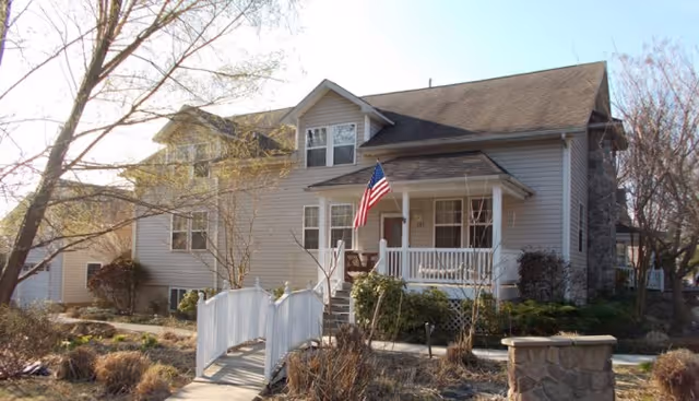 Exterior view of a two-story residential-style building with beige siding and a front porch displaying an American flag. A small white footbridge leads to the entrance, surrounded by leafless trees and dry landscaping under a clear sky.
