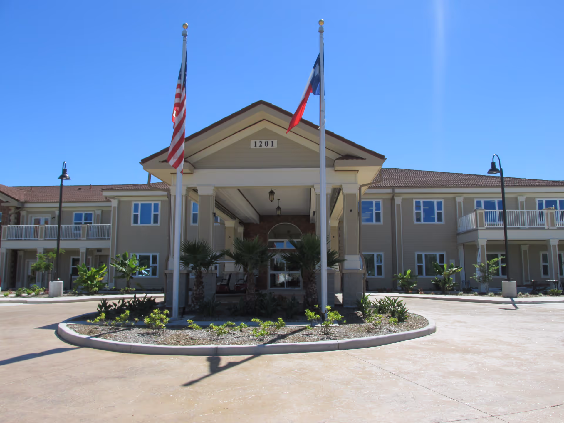 Front exterior view of The Gardens at Brook Ridge Assisted Living & Memory Care building with two flagpoles displaying the American and Texas flags, a covered entrance, and a circular driveway with landscaping under a clear blue sky.